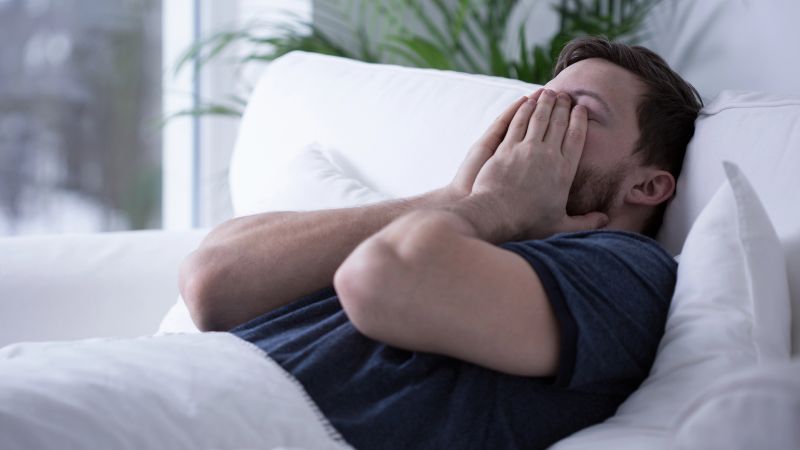man lies on a white couch covering his face with his hands