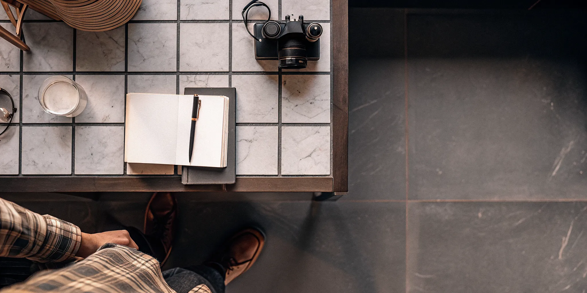 Person in plaid shirt sitting at a table with a camera, notebook, pen, and glass of water, reflecting a creative workspace atmosphere related to mental health treatment options.