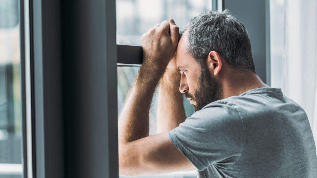 depressed bearded man leaning window looking